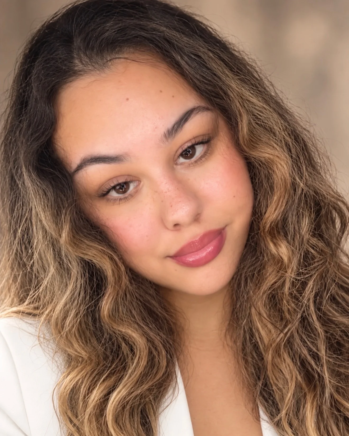 Arianna, esthetician at Lash L’Atelier, with long wavy honey-brown hair and a white blazer