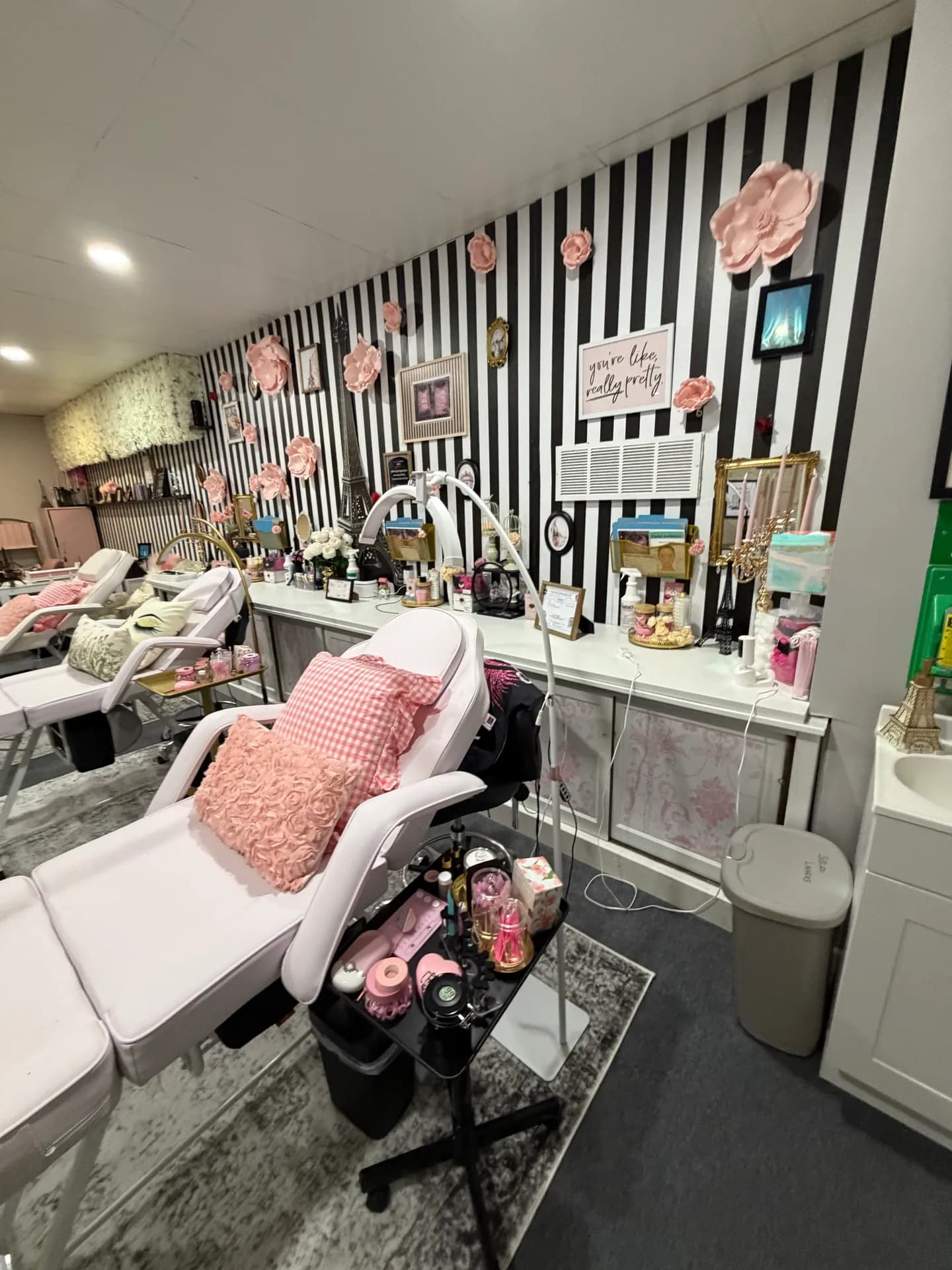 The lash bar: a white counter of lash tools, candlesticks and little Eiffel Towers along a black-and-white striped wall with pink paper flowers, beside a treatment bed with gingham and rose pillows