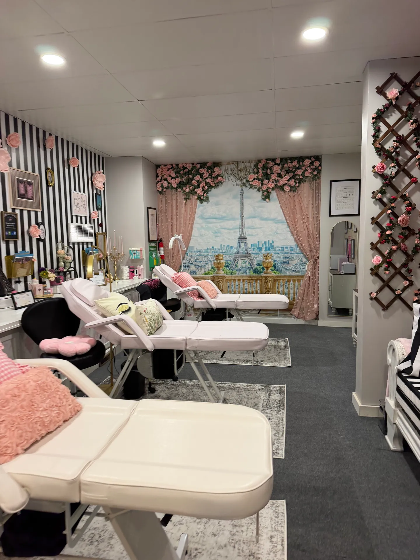 The Lash L’Atelier treatment room: blush-pink treatment beds on grey rugs beside a black-and-white striped wall hung with paper roses, facing a mural of the Eiffel Tower framed by pink curtains and roses
